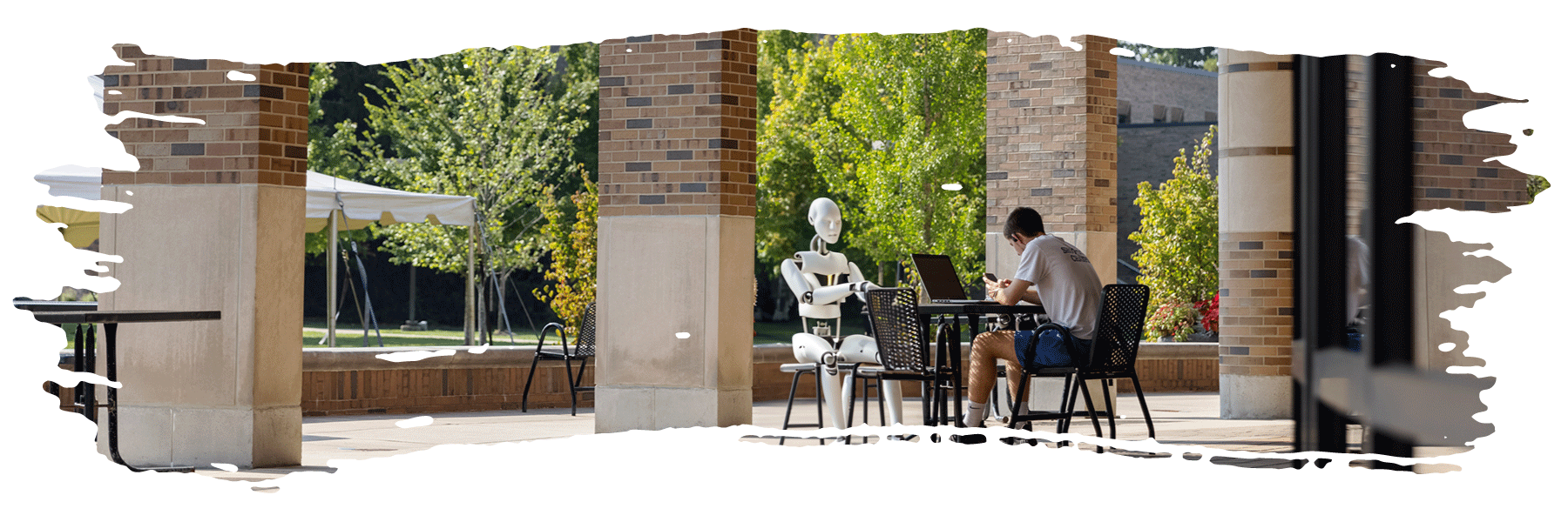 A student at his computer and a robot seated opposite him sit at a table outside a classroom building