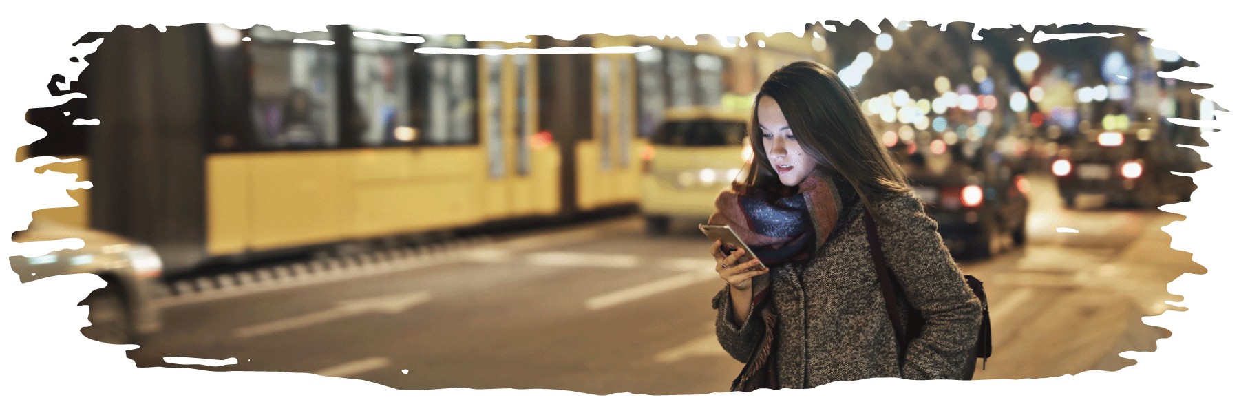 A woman is looking at her phone while standing on a busy street at nighttime with a winter coat on.