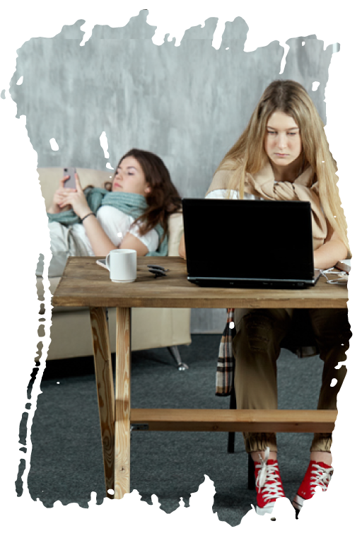 a college-aged woman working at her computer and another woman lying on a couch looking at her phone